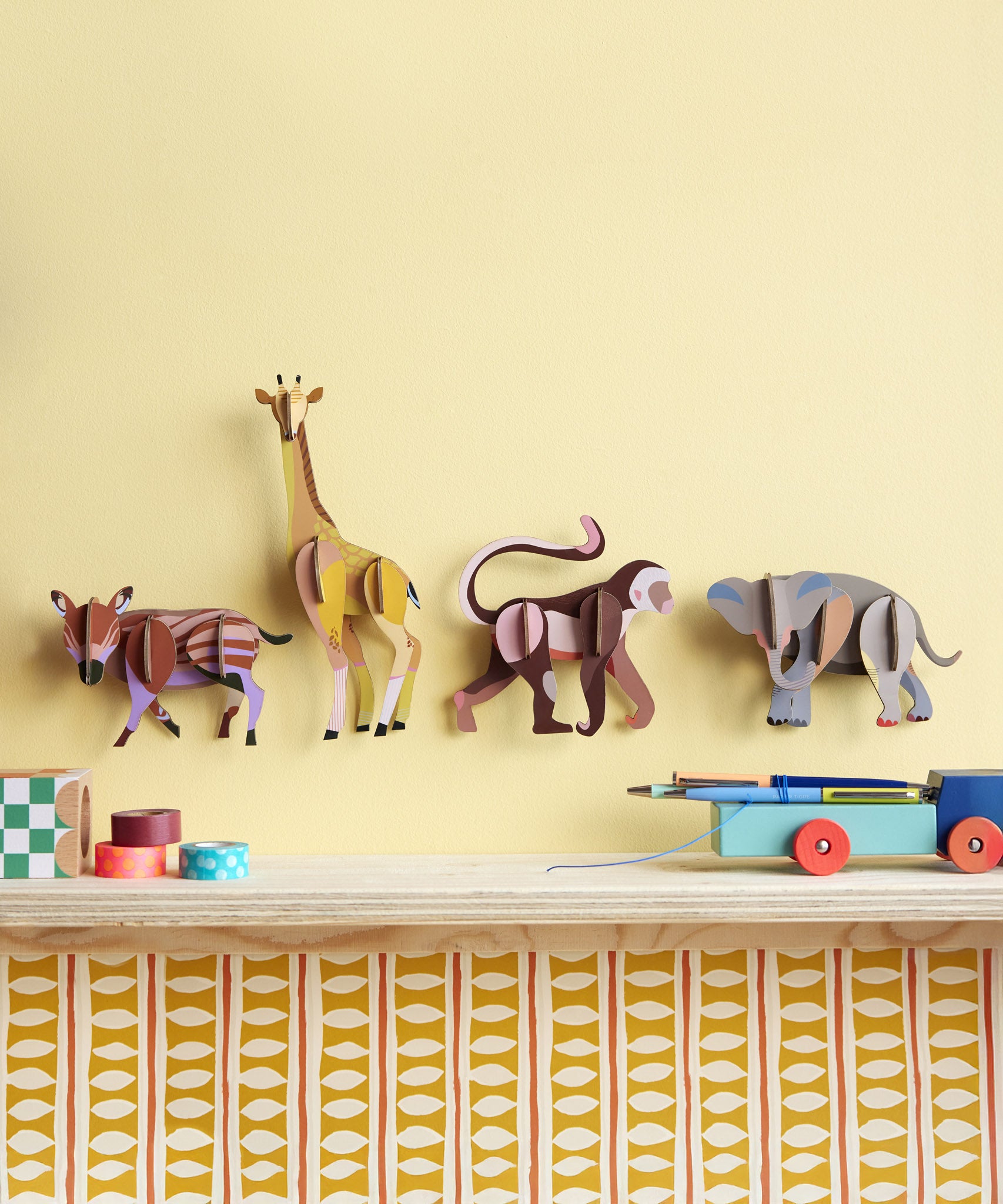 3 Studio Roof 3D Cardboard Models of Wildlife Animals on a yellow wall - the Boar Piglet, Giraffe, Monkey and the Elephant. They are surrounded by decorative objects.