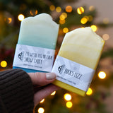 An adult hand holding the The Soapmine  handmade bucks fizz and snow fairy inspired fragrance bars available at Babipur in front of a festive background