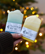 An adult hand holding the The Soapmine  handmade bucks fizz and snow fairy inspired fragrance bars available at Babipur in front of a festive background