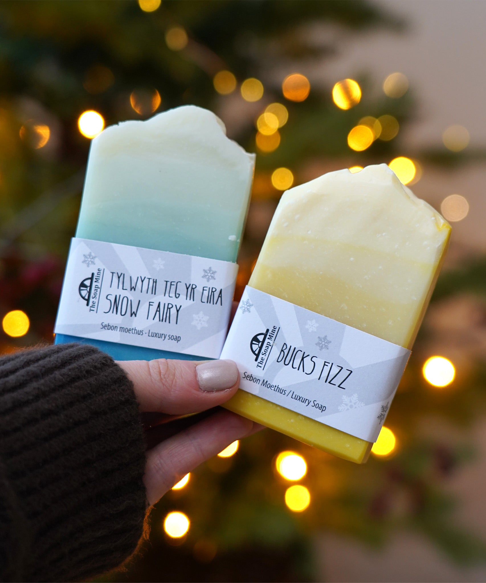 An adult hand holding the The Soapmine  handmade bucks fizz and snow fairy inspired fragrance bars available at Babipur in front of a festive background