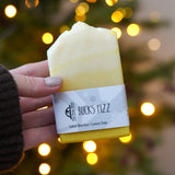 An adult hand holding the The Soapmine handmade bucks fizz bar available at Babipur in front of a festive background