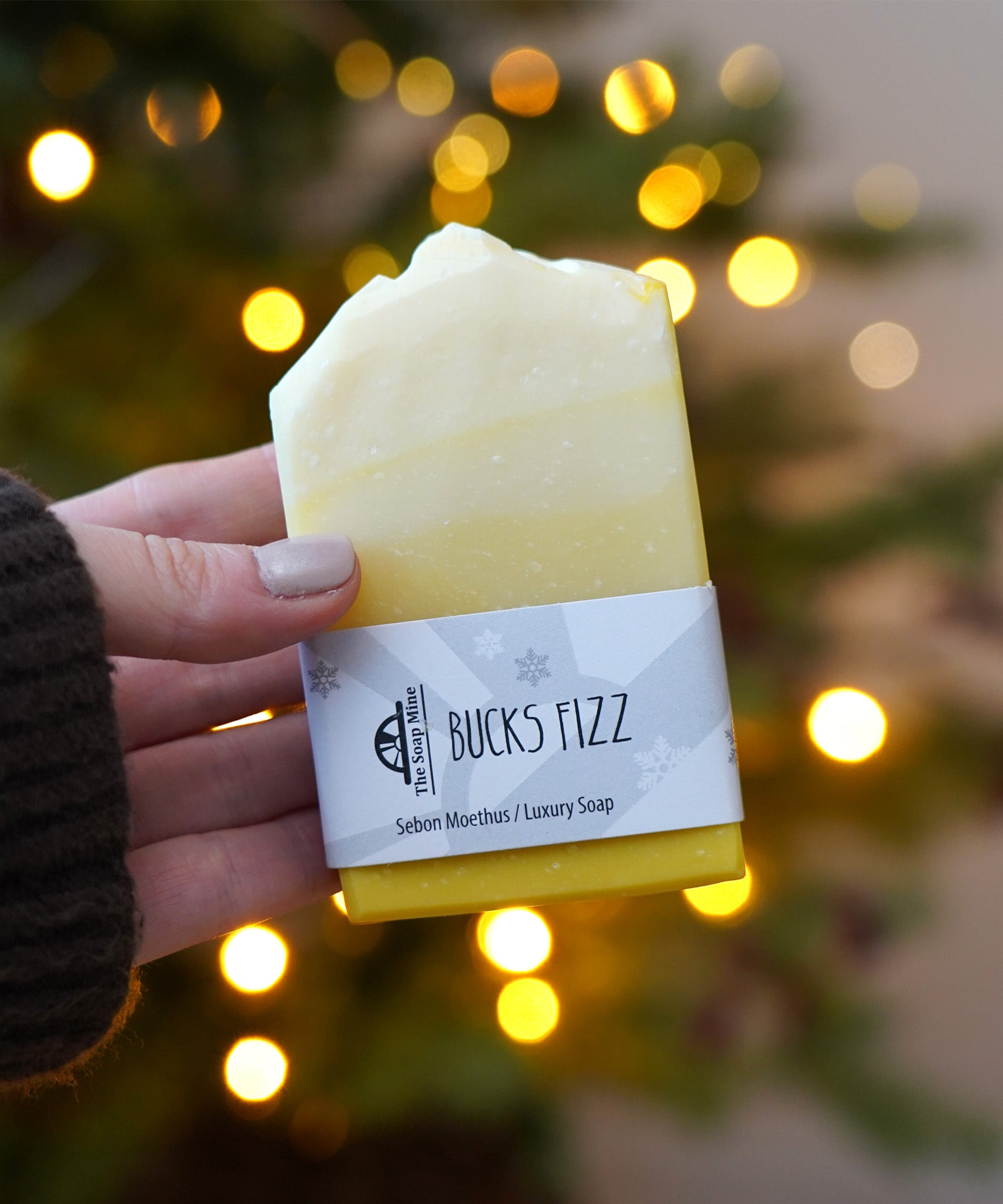 An adult hand holding the The Soapmine handmade bucks fizz bar available at Babipur in front of a festive background