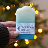 An adult hand holding the The Soapmine  handmade snow fairy bar available at Babipur in front of a festive background