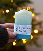 An adult hand holding the The Soapmine  handmade snow fairy bar available at Babipur in front of a festive background