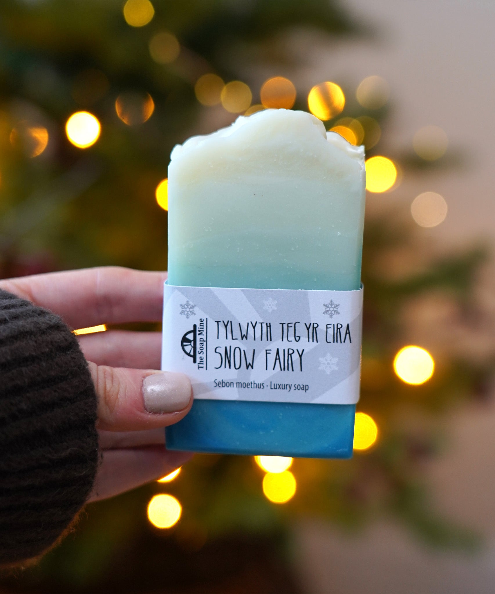 An adult hand holding the The Soapmine  handmade snow fairy bar available at Babipur in front of a festive background