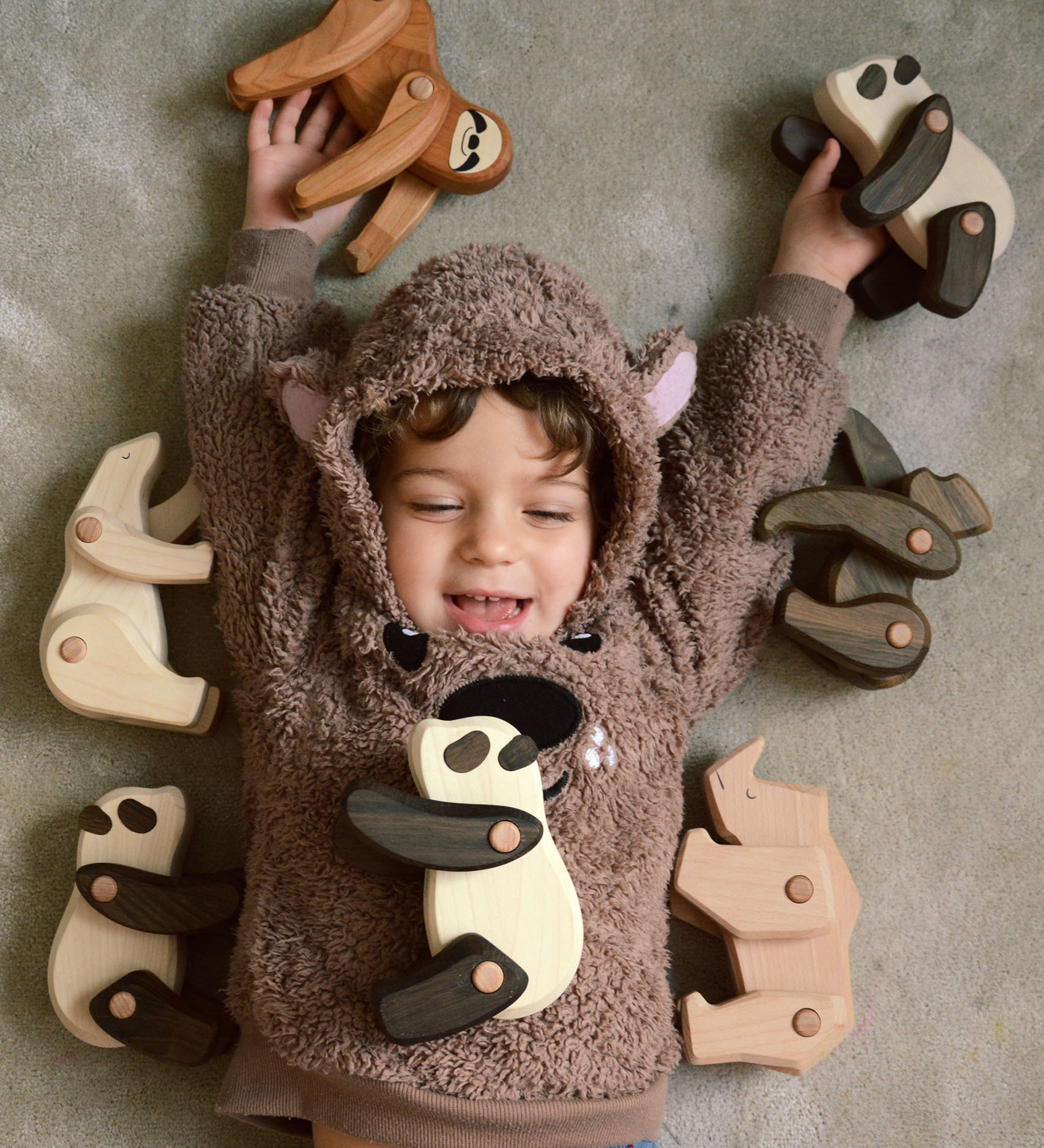 A child lying on the floor with a collection of the different Tobe wooden animals available at Babipur including a sloth, polar bear, panda, gorilla and a rhino.
