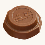 An angled view of the Tony's Chocolonely Tiny Mix chocolate button with the imprint of a broken chain on the front, to represent the breaking of slave labour. The image is on a cream background.