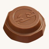 An angled view of the Tony's Chocolonely Tiny Mix chocolate button with the imprint of a broken chain on the front, to represent the breaking of slave labour. The image is on a cream background.