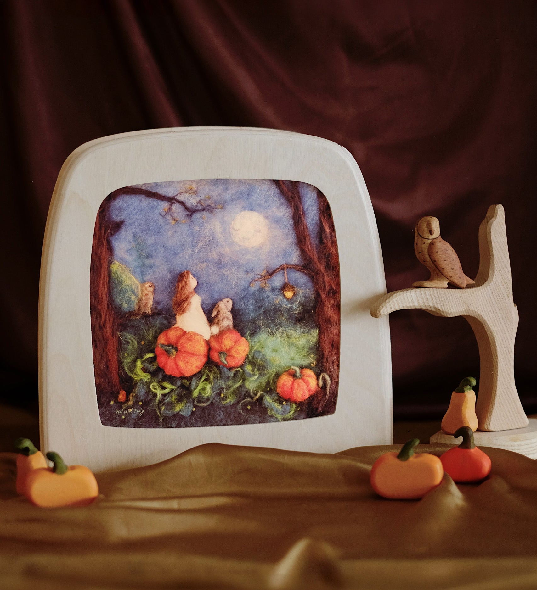 The Toverlux Magic Lamp with an illustration inside by Het Wol Feetje called Mother Moon. Next to the lamp is a natural wooden toy tree, with a wooden toy owl stood on one of the branches, and pumpkins  surround the tree and magic lamp. Everything is stood on a shiny gold cloth with a red cloth draped in the background