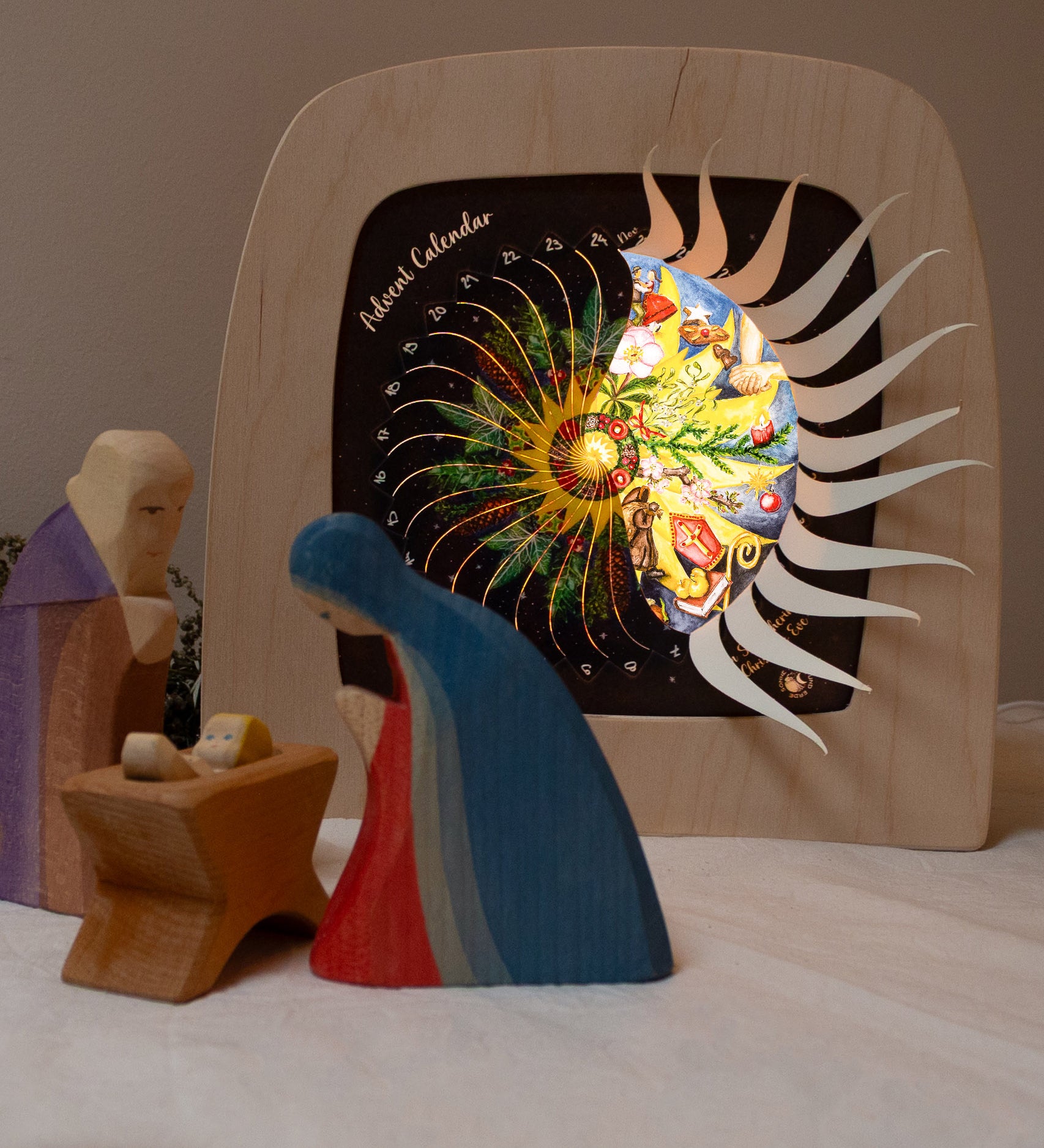 Mary and Joseph with baby Jesus wooden figures next to the Toverlux sonne mond und erde nativity advent calendar available at Babipur. Calendar has windows open revealing a lit up silhouette. 