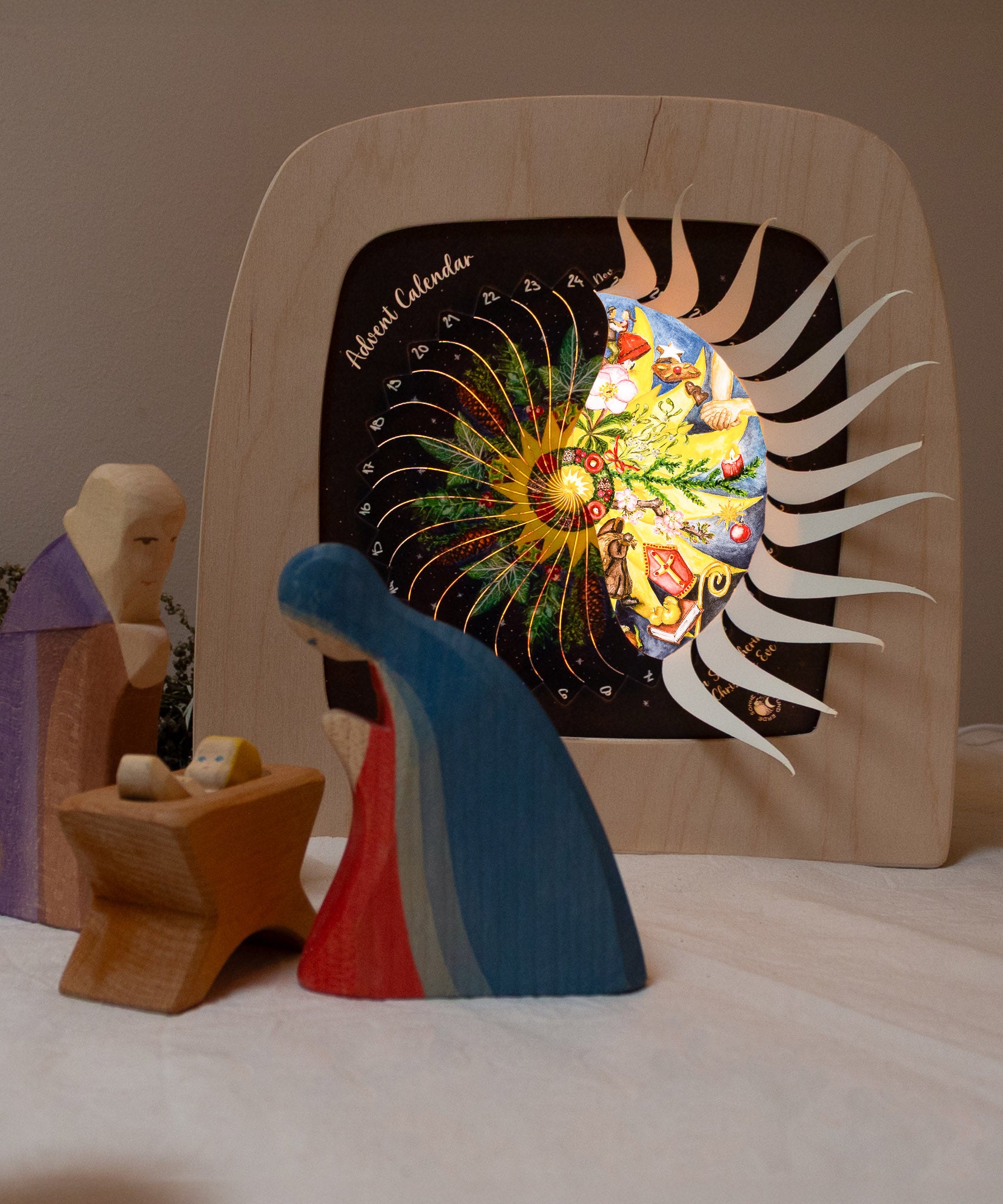 Mary and Joseph with baby Jesus wooden figures next to the Toverlux sonne mond und erde nativity advent calendar available at Babipur. Calendar has windows open revealing a lit up silhouette. 