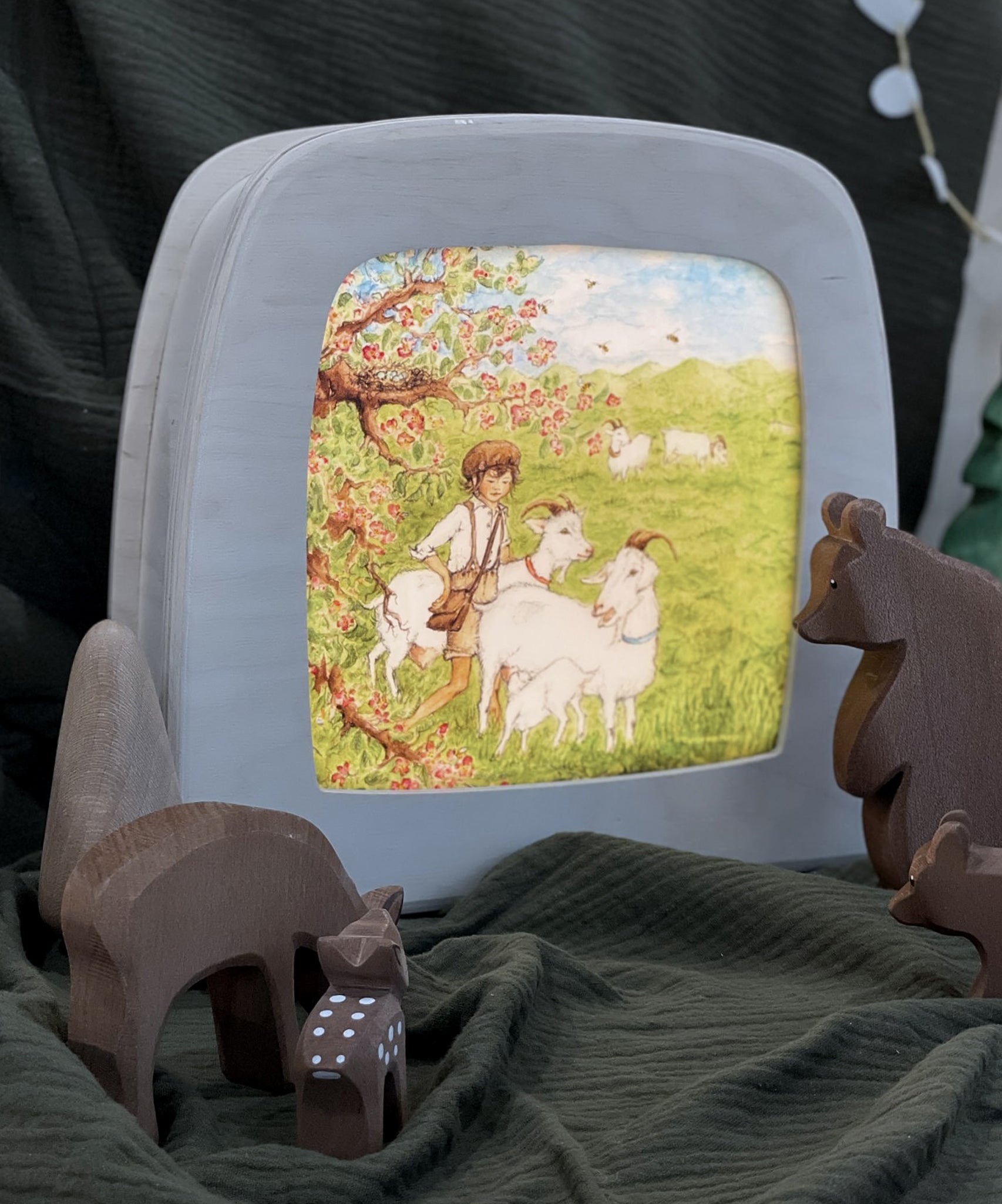 The Toverlux Magic Lamp with an illustration inside by Eentje Van Margo called Little Goat Herder. Looking at the Magic lamp are wooden, toy woodland animals with everything stood on green fabric and a green fabric background