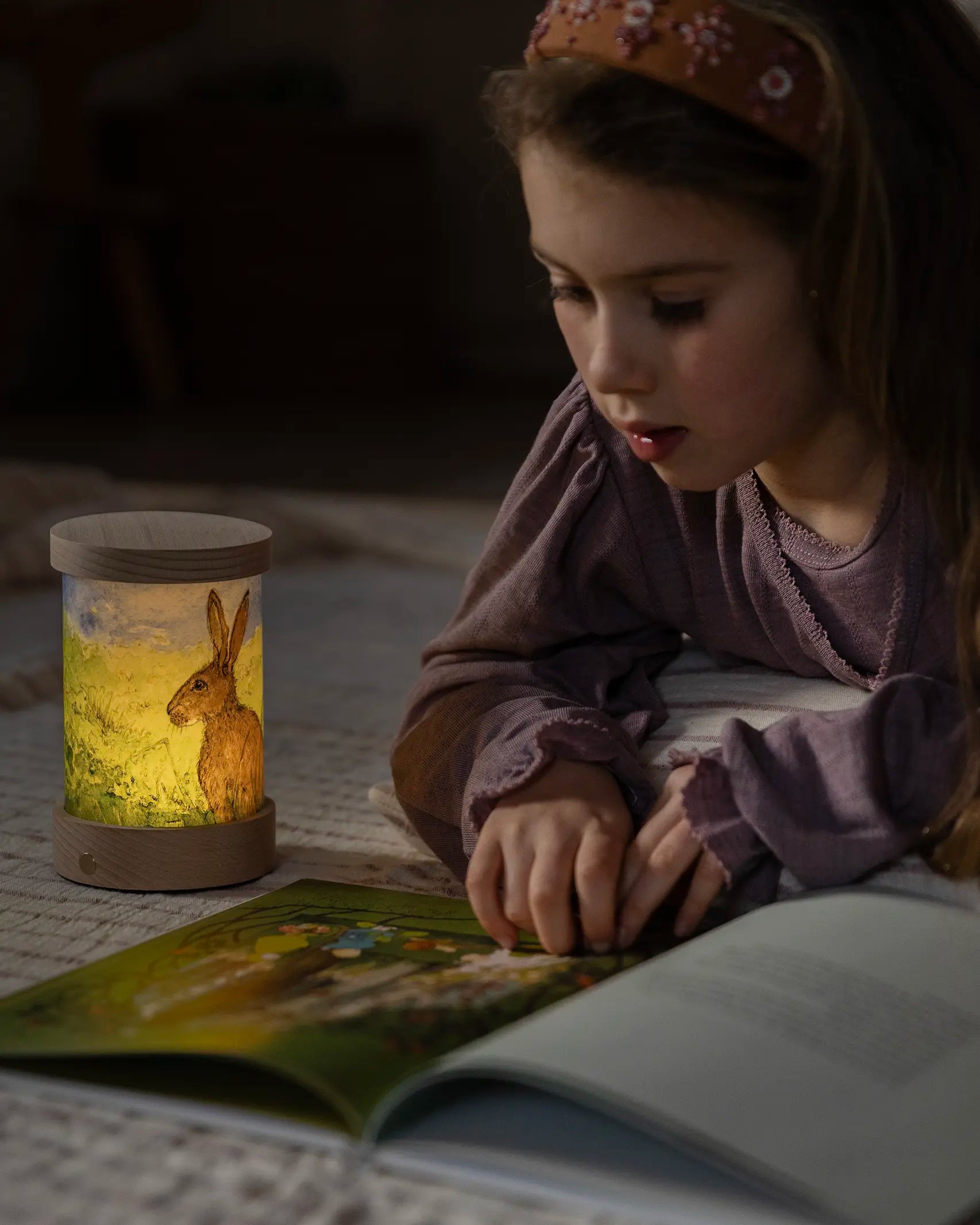 Child reading next to a Toverlux Wooden Lantern night light with rabbit artwork, it has a re-chargeable non-wired wooden base.