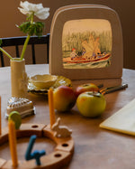 Toverlux Magic Lamp on table displaying artwork by Elsa Beskow, Peter in blueberry land, kids on a boat with a leaf sail.