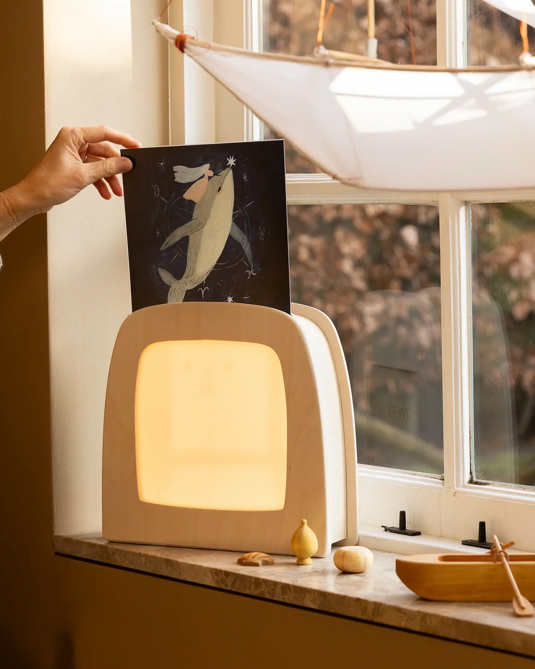 Hand placing a Toverlux Tijana Draws Whale silhouette into a Toverlux Magic Lamp Night Light Box on a windowsill.