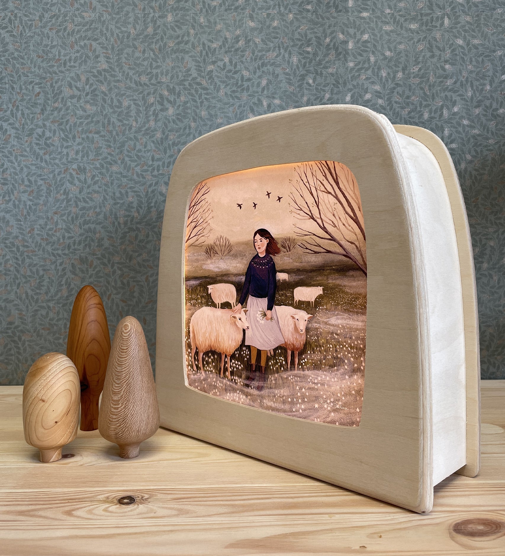 The Toverlux Magic Lamp with an illustration inside by Robin Pieterse called The Herdress. Next to the magic lamp are three wooden toy trees. The lamp and the tress are stood on a wooden table, with green patterned wallpaper in the background