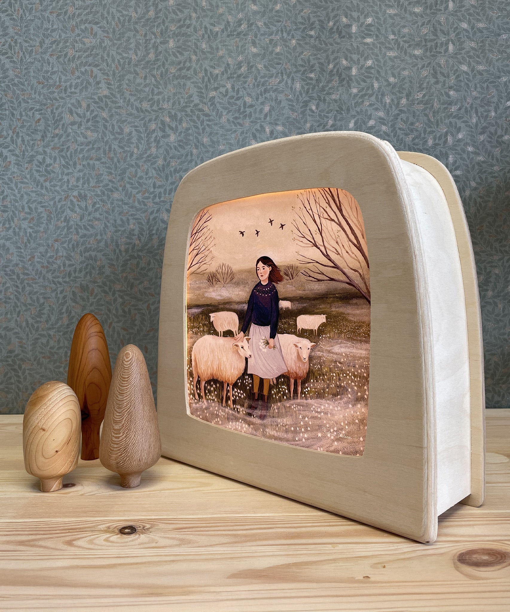 The Toverlux Magic Lamp with an illustration inside by Robin Pieterse called The Herdress. Next to the magic lamp are three wooden toy trees. The lamp and the tress are stood on a wooden table, with green patterned wallpaper in the background