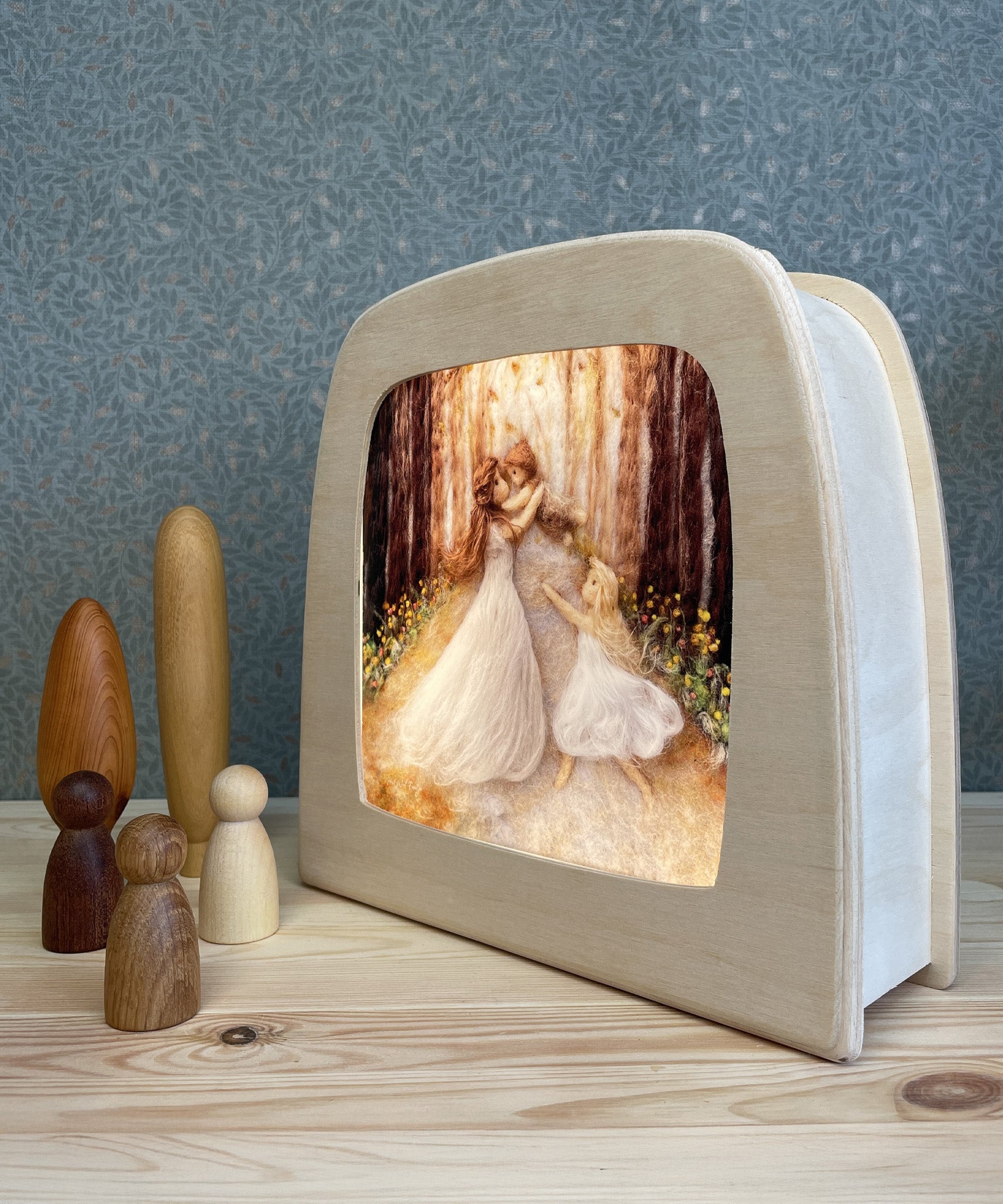 The Toverlux Magic Lamp with an illustration inside by Het Wol Feetje called Mothers Love. Next to the magic lamp are three wooden toy peg dolls and wooden toy trees. The lamp and the toys are stood on a wooden table, with green patterned wallpaper in the background
