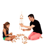 Father and daughter building a tower A using various wooden poles and blocks from the Trigonos mini large construction set