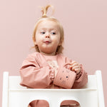 Baby sat in a high chair in a Trixie Animal Friends Waterproof Long Sleeve Bib, pink, Mrs Cat and matching sippy cup. 