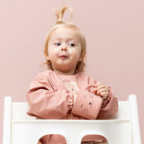 Baby sat in a high chair in a Trixie Animal Friends Waterproof Long Sleeve Bib, pink, Mrs Cat and matching sippy cup. 