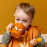 Baby drinking from the Trixie Animal Friends Silicone Sippy Cup, orange, Mr Fox. Wearing a matching bib. 