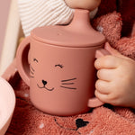 Child holding the handle of the Trixie Animal Friends Silicone Sippy Cup, pink, Mrs Cat.