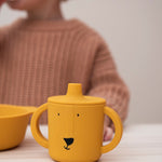 Trixie Animal Friends Silicone Sippy Cup, yellow, Mr Lion. Child wearing a pink waffle jumper in the background. 