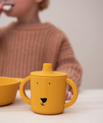 Trixie Animal Friends Silicone Sippy Cup, yellow, Mr Lion. Child wearing a pink waffle jumper in the background. 
