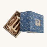 The Wooden Story Turtle Dove Rattle inside its cardboard box packaging. The rattle is laid on wood shavings