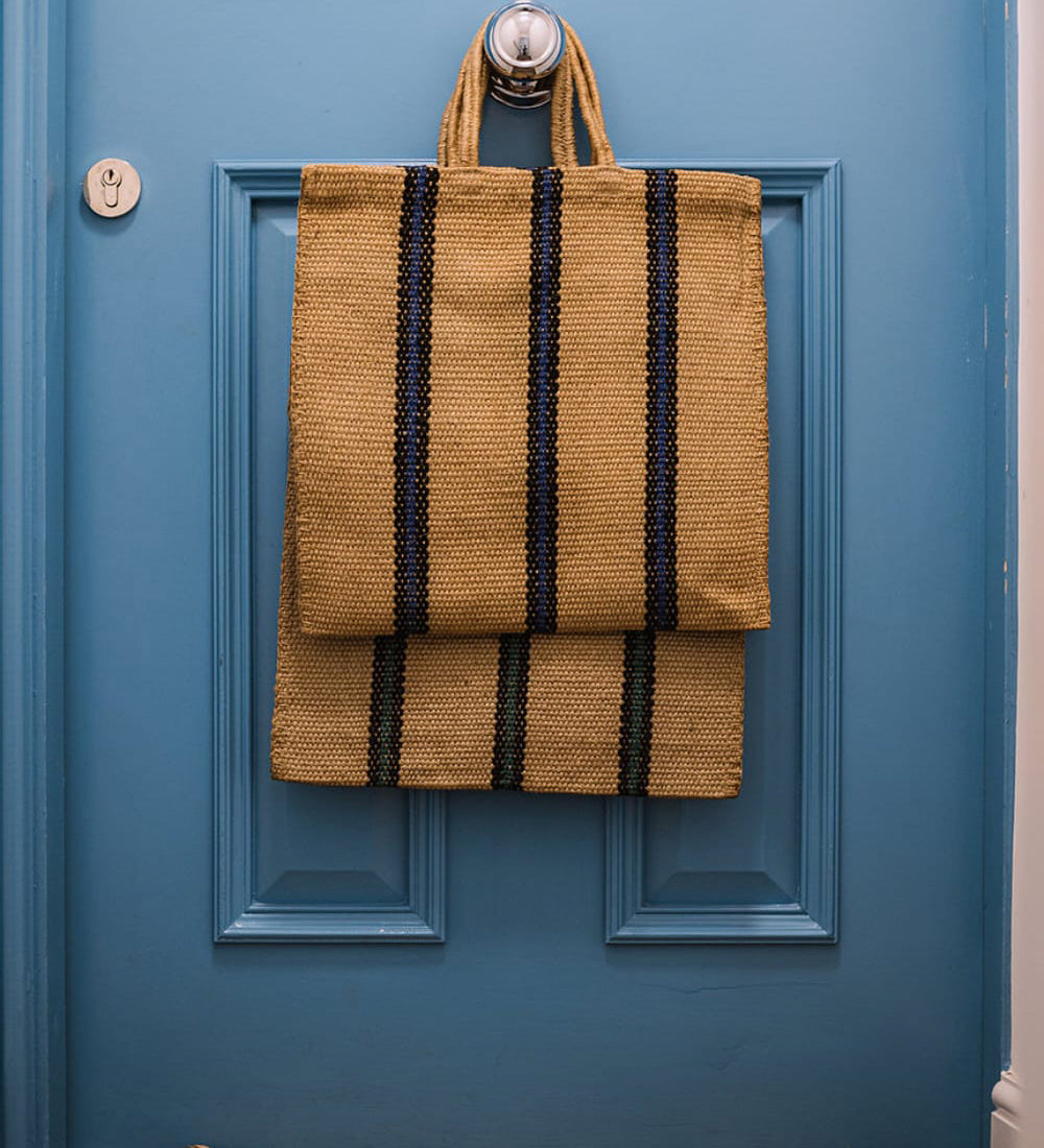 The Turtle Bags hand woven jute tote bag with short handles in a blue and black stripe design, hanging on the doorknob of a blue door