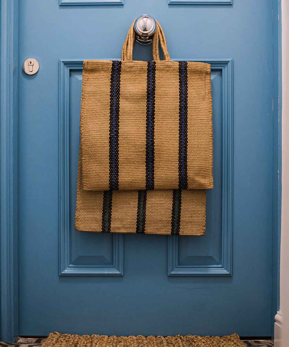 The Turtle Bags hand woven jute tote bag with short handles in a blue and black stripe design, hanging on the doorknob of a blue door