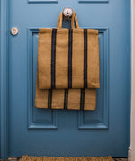 The Turtle Bags hand woven jute tote bag with short handles in a blue and black stripe design, hanging on the doorknob of a blue door