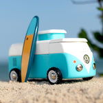 Candylab VW Beach bus in blue, with a blue and yellow surf board propped up next to the van, stood on a sandy beach with a blue sky background