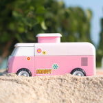 The side of the Candylab VW Beach bus in pink, stood on a sandy beach with green tree leaves and branches in the background. The image also shows colourful stickers on the side of the van