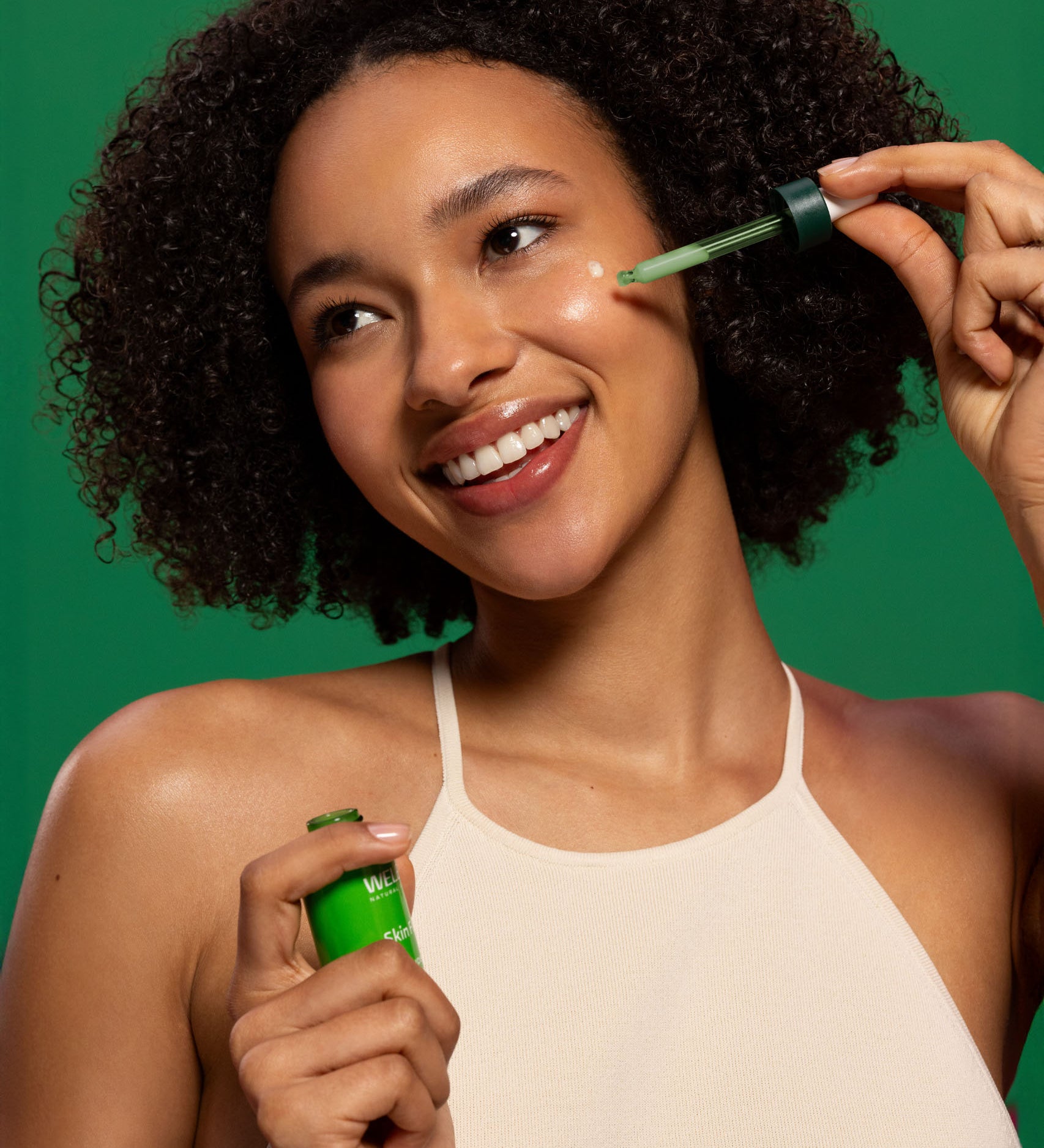 A woman applying the Weleda skin food super serum on her face and showing the  glass bottle and pipette. Weleda have a range of luxurious, natural skin care items available at Babipur. 