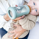 Toddler sitting on parents lap drinking from the Hevea Glass Bottle Wide Neck With Rubber Sleeve - 250ml in Seafoam Blue. 