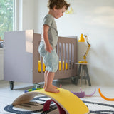 Wobbel original wooden balance board with a yellow felt backing, with child stood on top in a playroom setting.