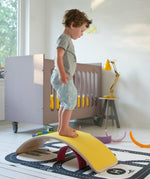 Wobbel original wooden balance board with a yellow felt backing, with child stood on top in a playroom setting.