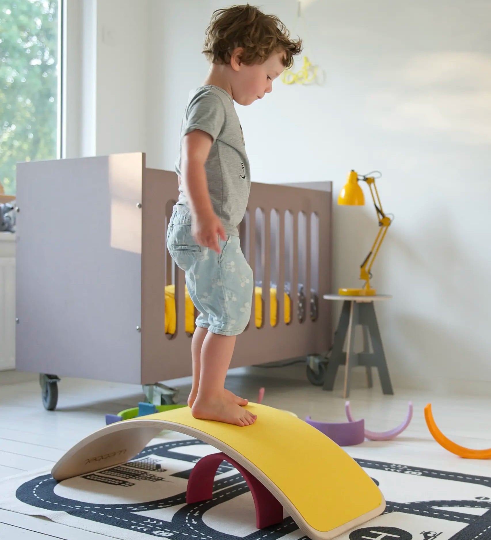 Wobbel original wooden balance board with a yellow felt backing, with child stood on top in a playroom setting.