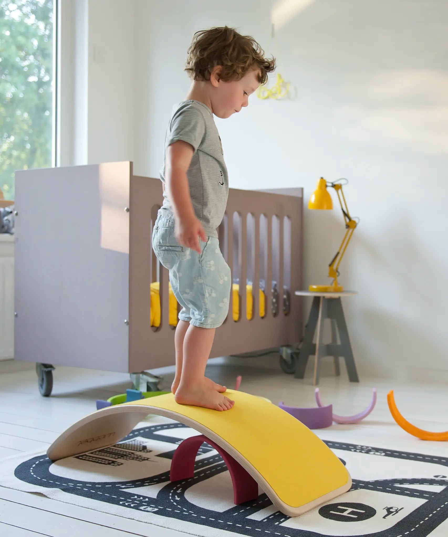 Wobbel original wooden balance board with a yellow felt backing, with child stood on top in a playroom setting.
