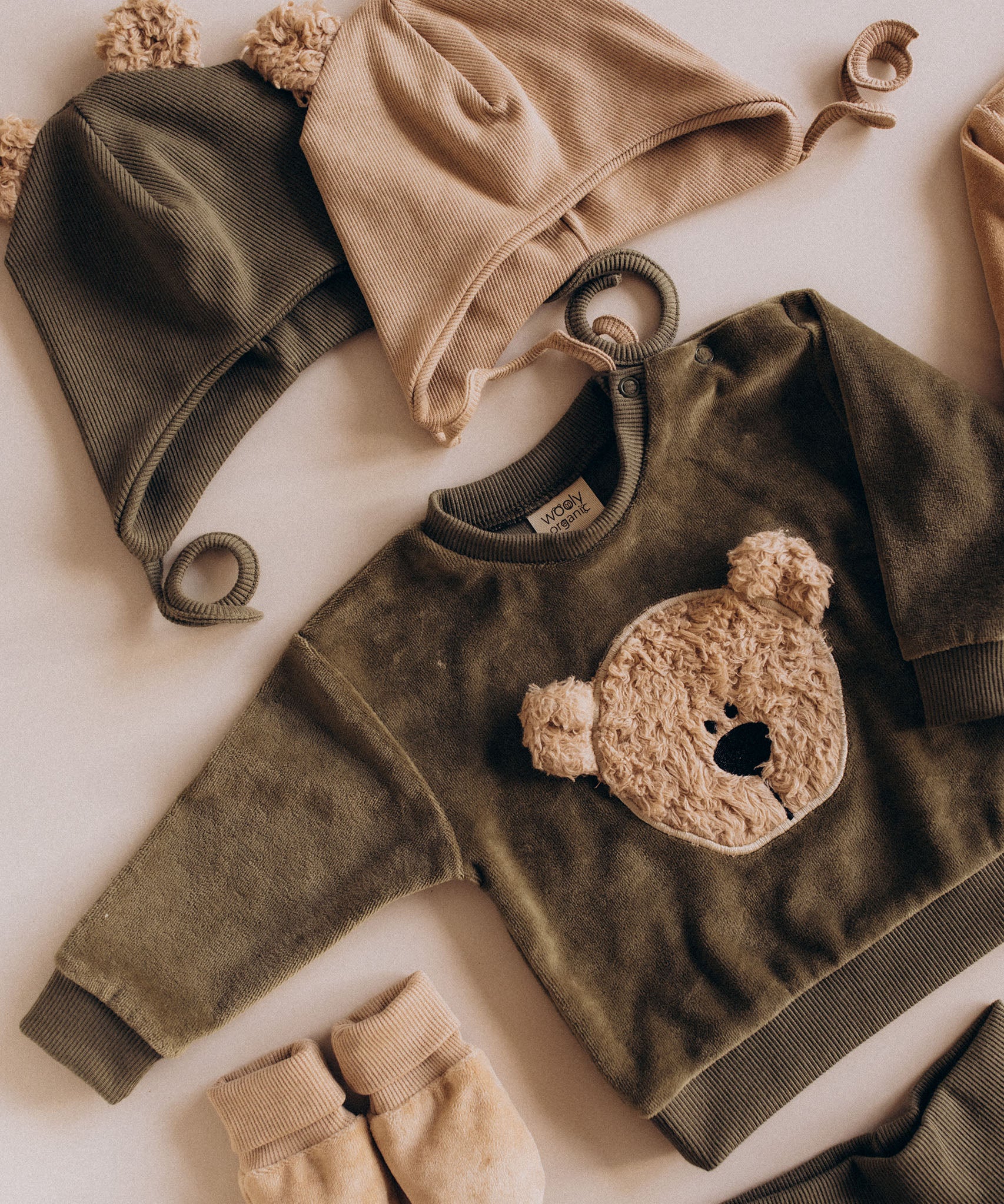 A flatlay showing the Wooly Organic embroidered sweater in tea leaf /savannah available at Babipur with the tea leaf and pale blush hat with teddy bear ears 