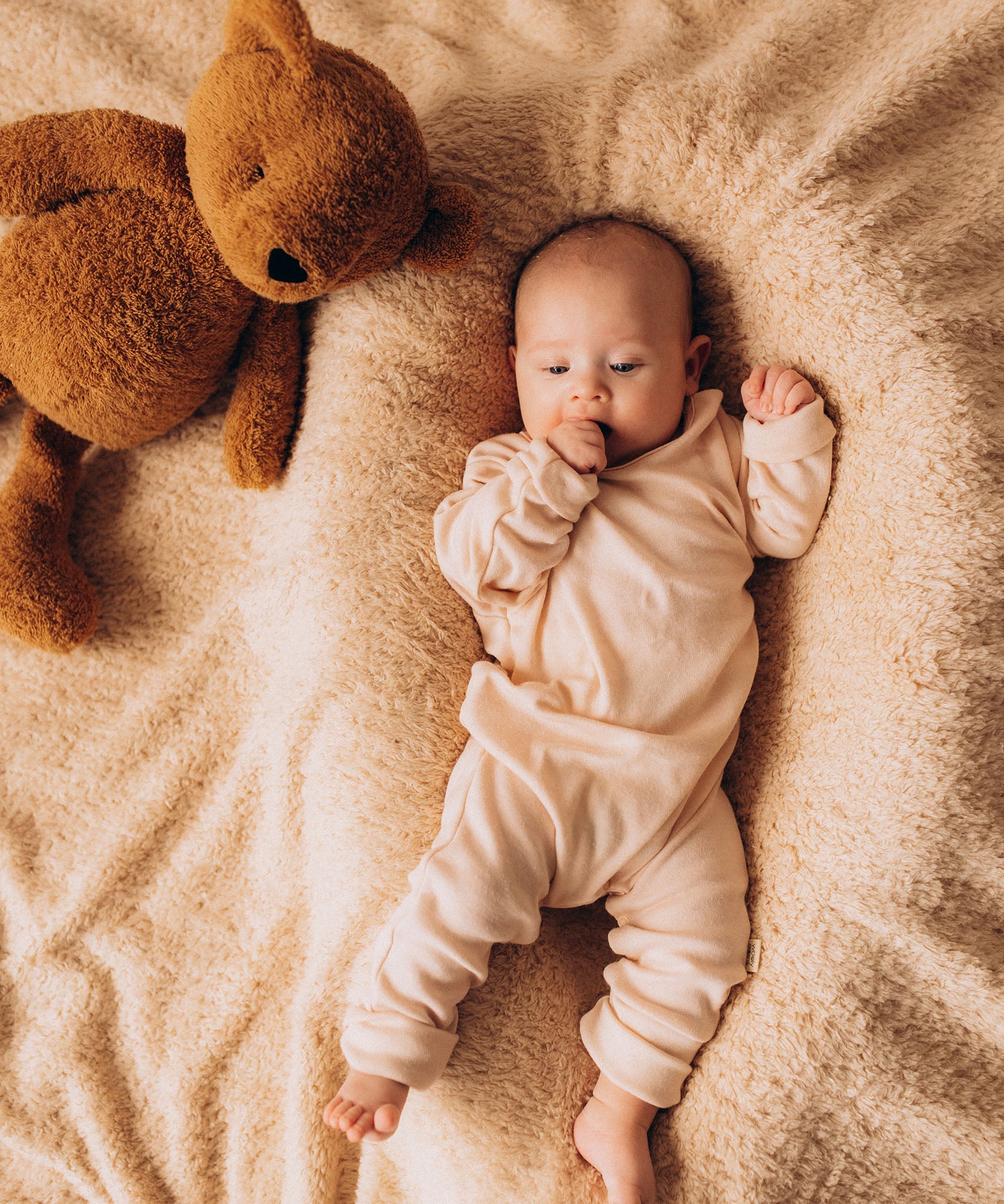 A top down image of a baby lying on their back next to the Wooly Organic soft teddy bear in caramel brown available at Babipur.