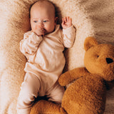 A close up of a baby lying on a soft and cuddly blanket next to the Wooly Organic soft teddy bear in caramel brown available at Babipur