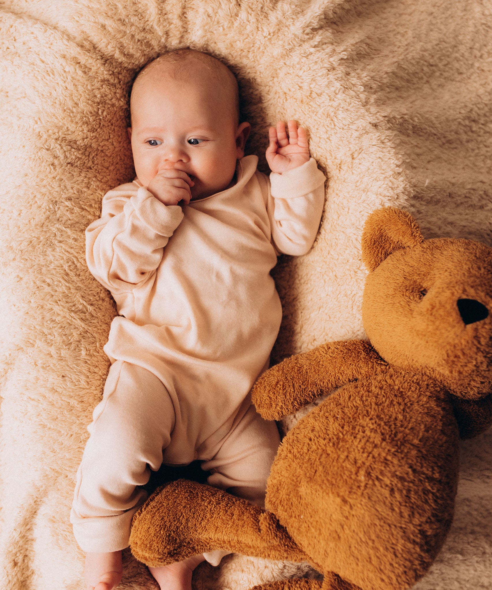 A close up of a baby lying on a soft and cuddly blanket next to the Wooly Organic soft teddy bear in caramel brown available at Babipur