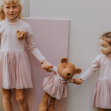 2 children standing next to eachother and holding the Wooly Organic soft teddy bear in caramel brown with a pink tutu by the hands. Teddy bear available at Babipur. 
