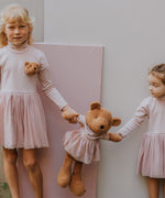 2 children standing next to eachother and holding the Wooly Organic soft teddy bear in caramel brown with a pink tutu by the hands. Teddy bear available at Babipur. 
