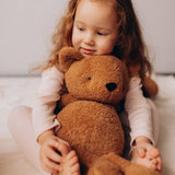 A close up of a small child holding the Wooly Organic soft teddy bear in caramel brown showing the cute facial features. Teddy bear  available at Babipur.