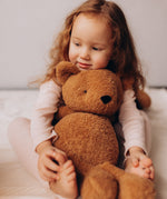 A close up of a small child holding the Wooly Organic soft teddy bear in caramel brown showing the cute facial features. Teddy bear  available at Babipur.