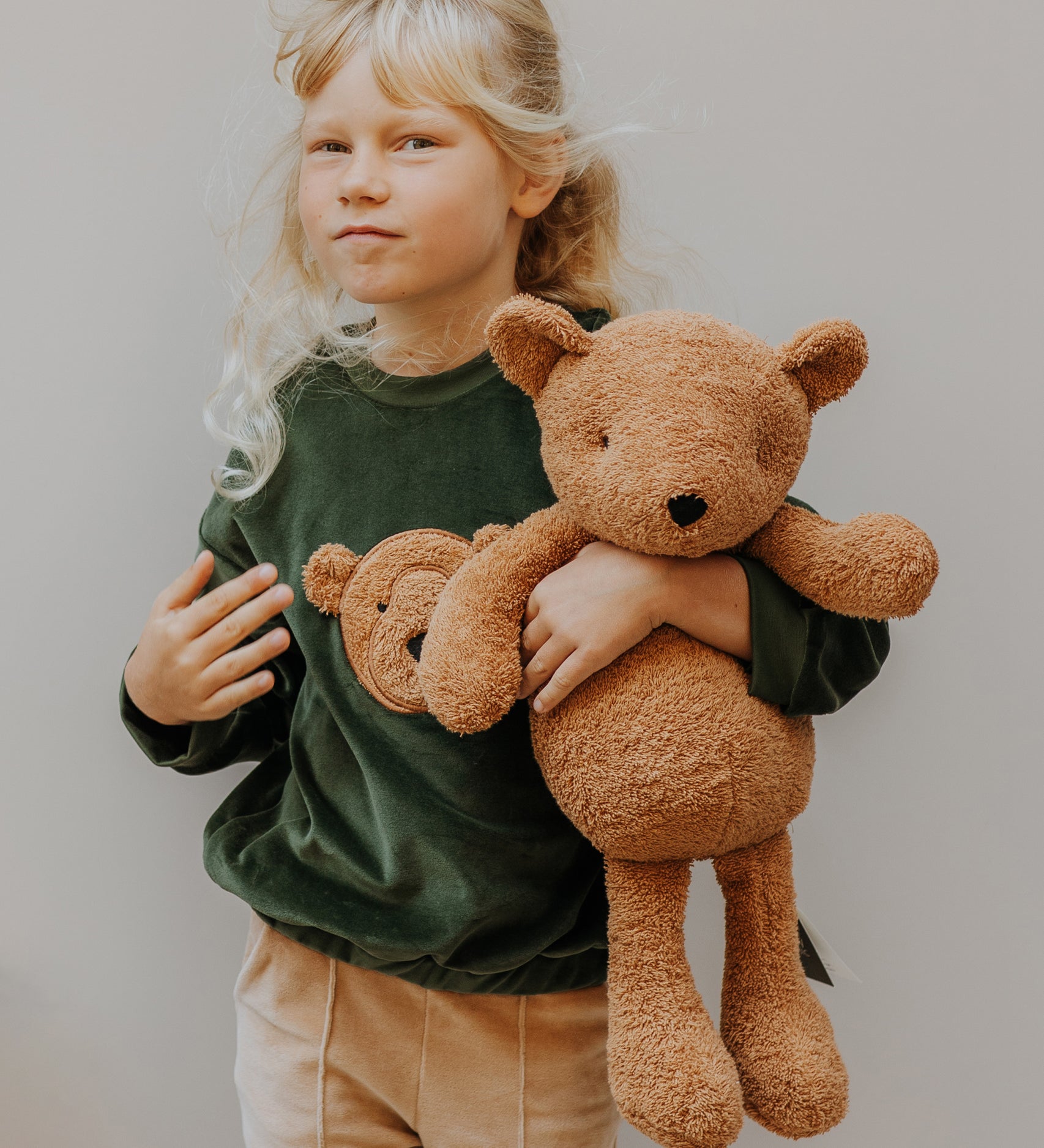 Child holding a large Wooly Organic teddy bear toy, made from organic cotton, in a caramel brown, for sale at Babipur.

